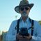 Der Künstler Pete Eckert mit seiner Mittelformatkamera vor blauem Himmel, er trägt Sonnenbrille und Hut