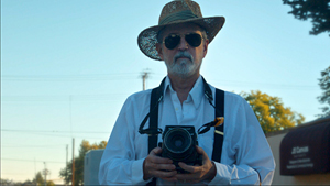 Der Künstler Pete Eckert mit seiner Mittelformatkamera vor blauem Himmel, er trägt Sonnenbrille und Hut